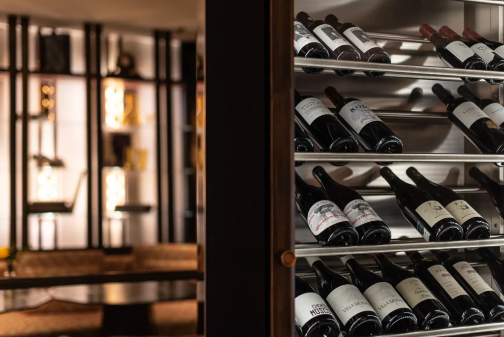 Wine bottles are stored on metal racks inside a wine cellar, with a softly lit lounge area in the background—perfect for enjoying drinks or pairing your favorite wine with food.